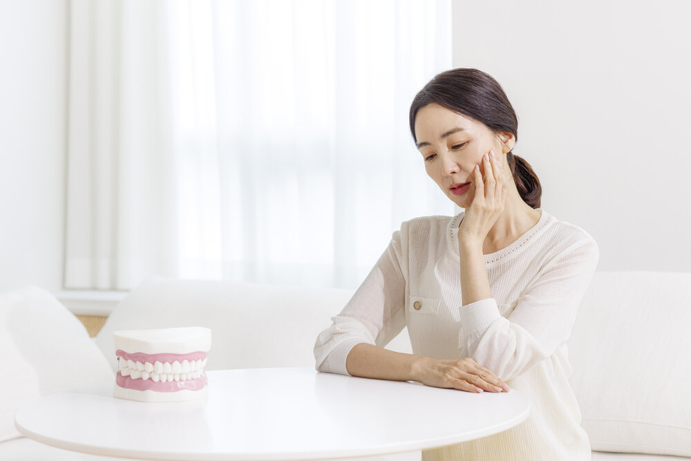 중년 여성이 테이블 앞에서 볼을 짚고 있고 옆에 치아 모형이 놓인 모습